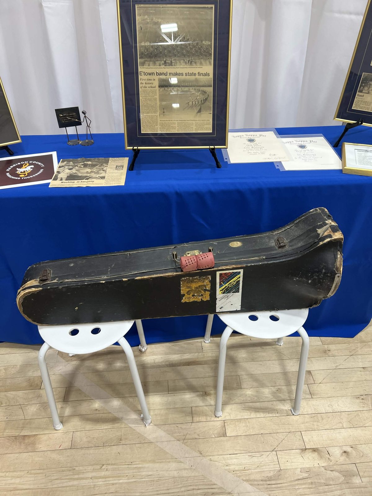 A battered black trombone case resting on two white stools in front of a table of framed newspaper clippings about the Elizabethtown band
